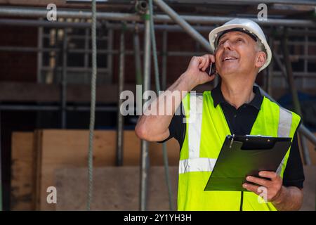Male builder foreman, construction worker, contractor or architect on building site writing on black clipboard and talking on cell phone Stock Photo