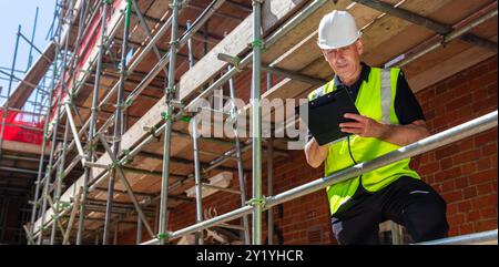 Male builder foreman, construction worker or site manager writing on a clipboard, wearing a white hard hat and hi vis vest Stock Photo