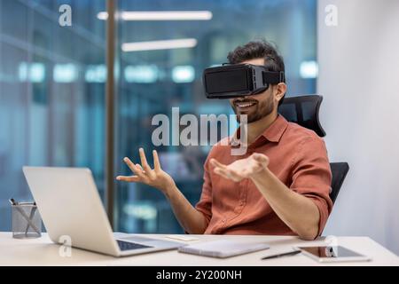 Business professional in modern office using virtual reality headset during video call. Engaged in immersive experience seated with laptop. Concept technology, innovation, and digital communication Stock Photo