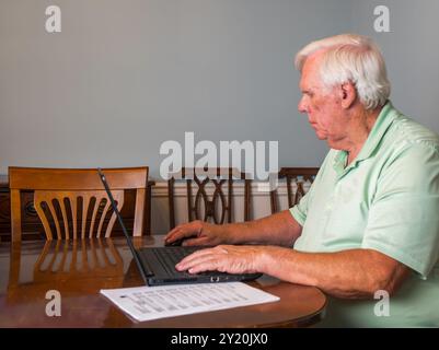 Older man with white hair and wearing a green shirt works on his laptop computer in a makeshift office in his dining room. Stock Photo