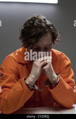 Pensive man wearing orange jumpsuit and handcuffs sitting alone at table, resting chin on hands, appearing thoughtful and focused in brightly lit room Stock Photo