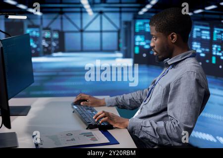 African american man typing on PC in server farm, ensuring data remains shielded from potential threats. IT expert coding at computer desk, safeguarding data center hardware against malicious access Stock Photo