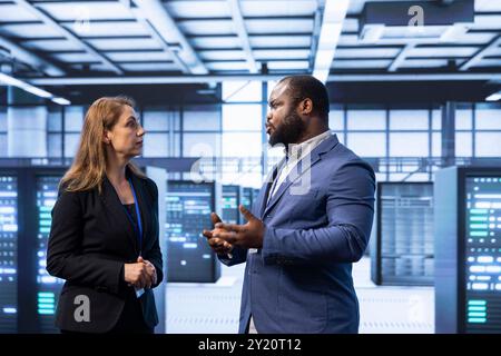 Colleagues doing brainstorming in server hub after installing storage units. IT staff member talking in data center, doing maintenance after replacing mainframes components Stock Photo