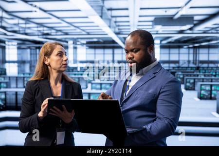 Coworkers in data center using notebook to identify and solve technical issues related to data center infrastructure. Team of admins managing, maintaining and securing server farm environment Stock Photo