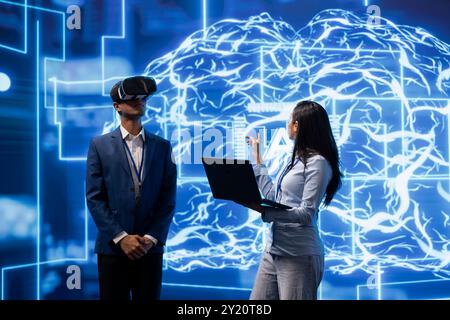 Engineer wearing VR headset looks at hologram of artificial intelligence human brain simulation, assisted by colleague using laptop. Team of developers interacting with AI visualization Stock Photo