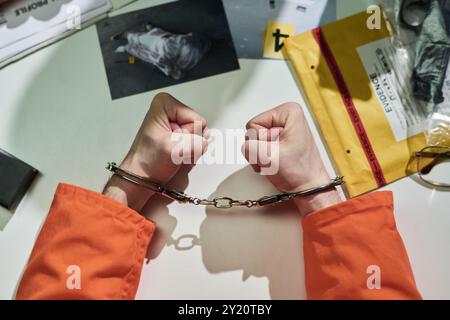 Handcuffed hands wearing orange jumpsuit resting on table with evidence envelopes, photos, documents, and handcuffs creating an investigative atmosphere Stock Photo