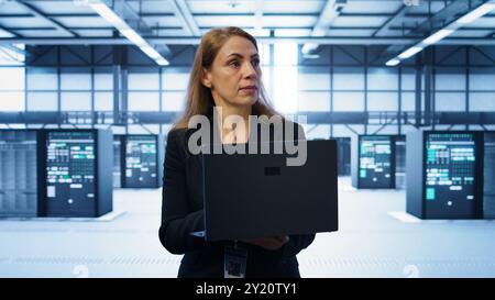 Software developer walking in data center, overseeing server operations using laptop. Woman ensuring optimal performance and data integrity in server farm using notebook, camera B Stock Photo