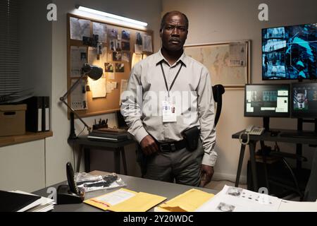 Portrait of African American FBI agent wearing badge, standing in a dimly lit office with various screens and bulletin board in background examining documents Stock Photo