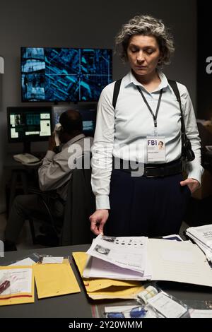 Gray-haired woman with FBI badge reviewing documents at office desk, team member analyzing data on monitors in background Stock Photo