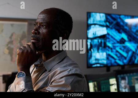 Elderly African American man thinking deeply in modern office filled with advanced technology and large screens depicting data. Hand on chin indicating contemplation and analysis Stock Photo