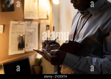 Man taking notes in office while reviewing documents on wall with focused expression and office lit warmly by desk lamp Stock Photo