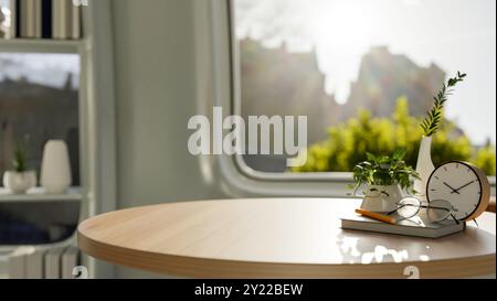 A copy space for displaying products on a round wooden table features eyeglasses, a book, a pencil, a clock, and decorative plants on the table in a m Stock Photo