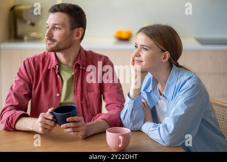 Smiling young couple staring into the distance while having tea and breakfast together in kitchen Stock Photo