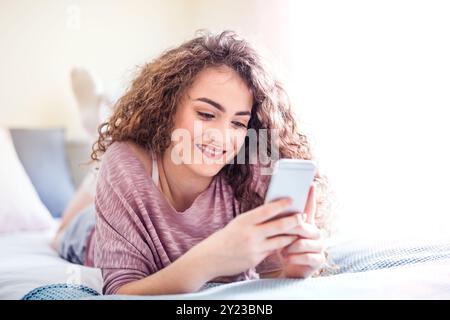 Beautiful young woman lying on her stomach on bed, scrolling on smartphone, smiling. Stock Photo