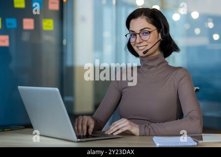 Smiling woman with glasses and headset uses laptop in office setting. Engaged in online meeting or support call. Modern technology enhances communication and productivity in business environment. Stock Photo