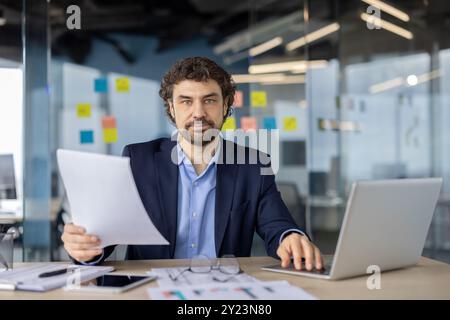 Serious businessman working in modern office holding document. Thoughtful professional analyzing information while using laptop. Focused on paperwork, enhancing productivity Stock Photo