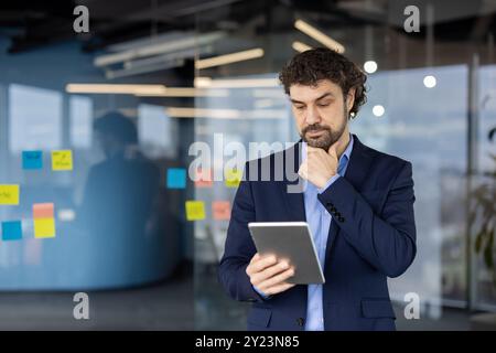 Businessman in blue suit deeply analyzes information on tablet. Thoughtful expression highlights focus on data-driven decision making in modern office setting with colorful sticky notes. Stock Photo