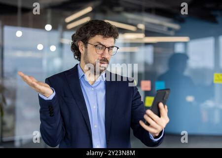 Businessman in blue suit looks confused while receiving bad news on smartphone in modern office. Expressive gesture conveys frustration and concern. Stock Photo