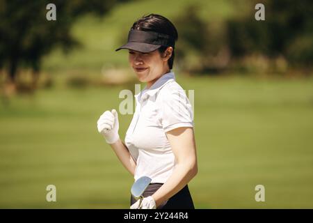 Empowered female Asian golfer in a cap and golf gloves exudes happiness and confidence while enjoying a sunny day on the golf course. Stock Photo