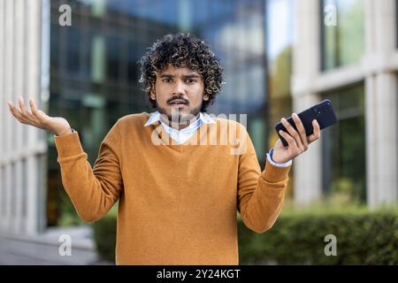 Man with curly hair in sweater holding smartphone with confused expression. Outdoor urban background suggests business context, conveying uncertainty, doubt, or perplexity in modern communication. Stock Photo
