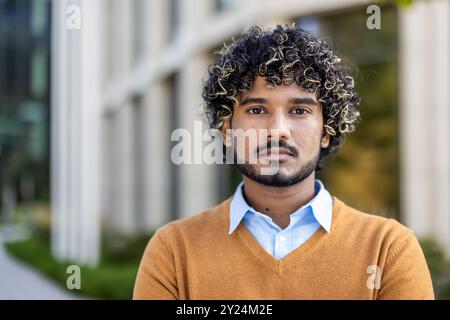 Portrait of confident young man with curly hair wearing a sweater and shirt. Standing outdoors in urban setting, looking towards camera with serious expression. Captures professional mood Stock Photo