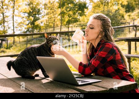 Young woman enjoys a peaceful break in the countryside, working remotely on her laptop in a park, surrounded by nature, petting her cat and sipping a Stock Photo