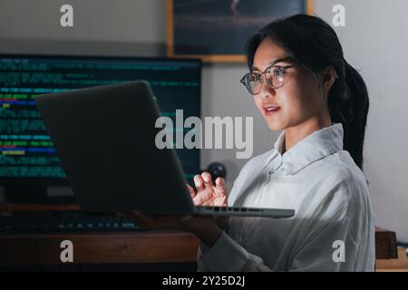 A young woman with glasses is working on a laptop in a dimly lit room. She appears focused and engaged, with a computer screen displaying code in the Stock Photo