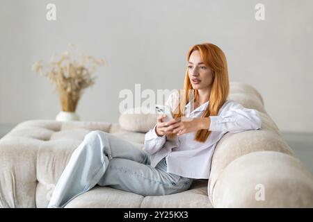 Attractive smiling young business woman relaxing on a leather couch at home, holding mobile phone Stock Photo