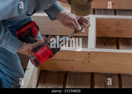 The hands of a man in his 80s on his knees bending over a framed wall changing the drill bit on his red Craftsman drill, USA. Stock Photo