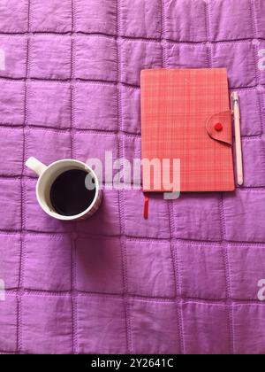 Vertical Directly Above Shot of a Vibrant Pink Eco-Leather Notebook, Pen, and Coffee on a Purple Blanket Stock Photo