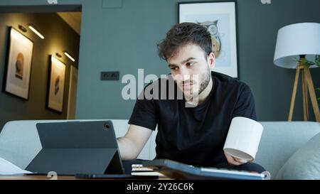 A man sits on a couch, holding a coffee mug while focusing on his tablet. Reviewing important work or studies. Stock Photo