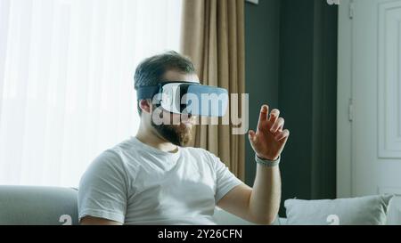 A man in a white shirt is seated on a couch, wearing a VR headset and reaching out as he interacts with the virtual world. The immersive nature of cut Stock Photo