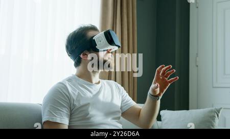 A man in a white t-shirt wearing a VR headset is fully immersed in a virtual reality experience, extending his hand as he interacts with the digital e Stock Photo