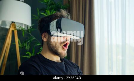 A young man experiences virtual reality with a VR headset in a living room. Amazement and excitement as he explores immersive digital worlds. Stock Photo
