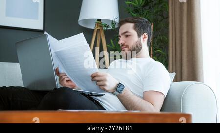 A man laying on a couch reviews financial documents while using a laptop. Focus and responsibility involved in managing personal finances, bills, and Stock Photo