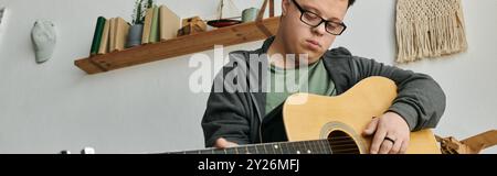 Young man with Down syndrome enjoys playing guitar surrounded by warm home decor. Stock Photo