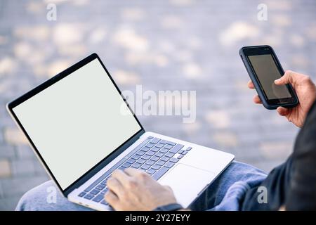 A person using a laptop and a smartphone simultaneously while sitting outdoors. The laptop has a blank screen, and the individual is typing on the key Stock Photo