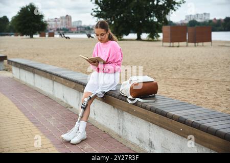 A young woman with a prosthetic leg relaxes outdoors, reading a book by the calming waterfront. Stock Photo