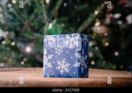 A small square present wrapped in blue snowflake wrapping paper with Christmas tree in background Stock Photo
