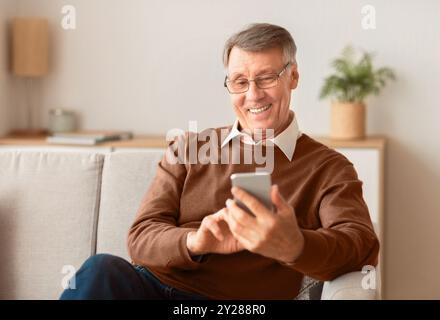 Joyful Elderly Man Using Cellphone Sitting On Sofa At Home Stock Photo