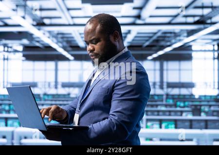 Computer scientist using laptop in data center with advanced tech, ensuring cybersecurity and efficient network management. African american expert in server hub optimizing data center with device Stock Photo