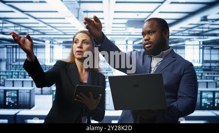 Teamworking technicians managing data center with advanced technology using devices. Coworkers in server room optimizing data center infrastructure with laptop and tablet, camera B Stock Photo