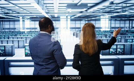 Data center engineer presenting equipment to inspector supervising operations. IT specialist in server farm doing tour for supervisor monitoring equipment performance, camera B Stock Photo