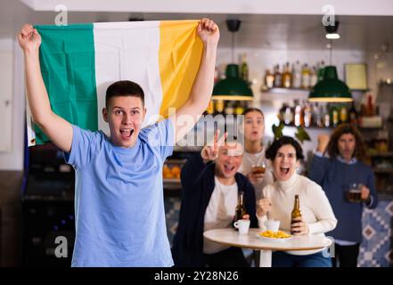 Company of emotional young adult fans supporting favorite team with flag of Ireland while resting in pub with beer Stock Photo