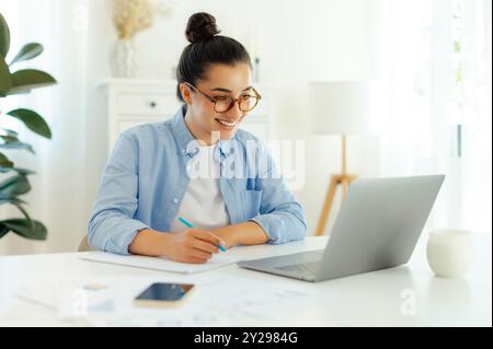 Concentrated successful mixed race brunette woman in a blue shirt, sits at a work desk while working in the laptop, doing online tasks, develop new digital project, planning marketing strategy, smiles Stock Photo