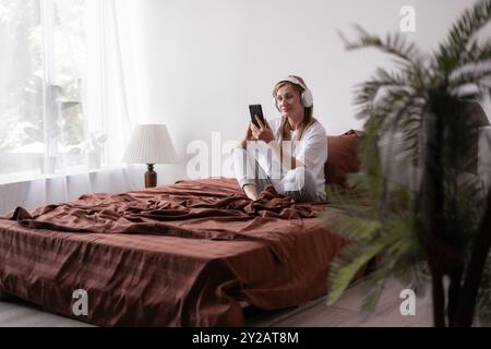 Cheerful older woman in headphones looks at smartphone, lies on brown bed listen favorite music. Relax, rest and lifestyle Stock Photo