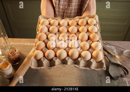 A man holds a large egg carton, with 30 eggs visible in it. In the background is a kitchen setting with utensils and jars. The focus is on the eggs in Stock Photo