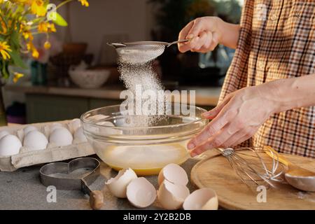 Close-up of female hands sifting flour and preparing dough in a transparent bowl against the background of a kitchen. Eggs in a box and a tool for mak Stock Photo