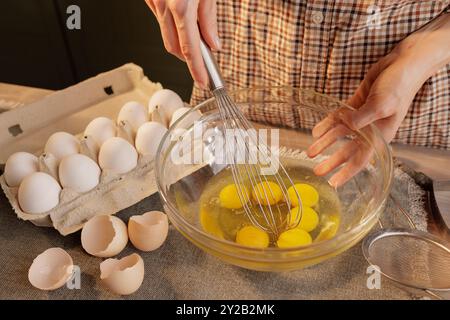 Close-up of female hands beating eggs with a hand whisk in a transparent bowl against the background of a kitchen table. Eggs in a box and a tool for Stock Photo
