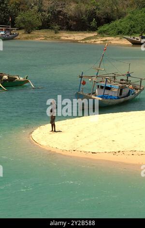 A man standing on sandy beach in a background of fishing boats on a lagoon-like seascape in the fishing beach of Pero in Sumba Island, Indonesia. Stock Photo